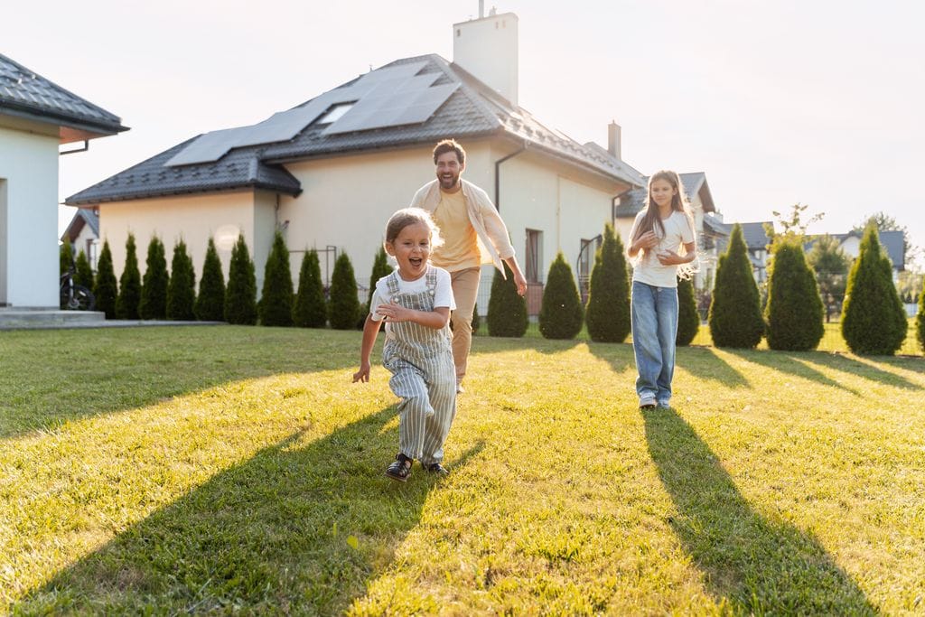 Family running and playing on a sunlit lawn in front of a suburban home with rooftop solar panels, highlighting clean energy and everyday home life.