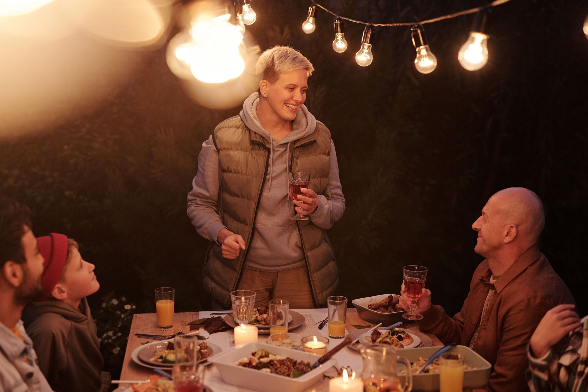A family gathers around an outdoor dinner table at dusk, warmly lit by hanging string lights. A smiling woman with short blonde hair stands holding a glass, raising a toast with an older man across the table while two others look on.
