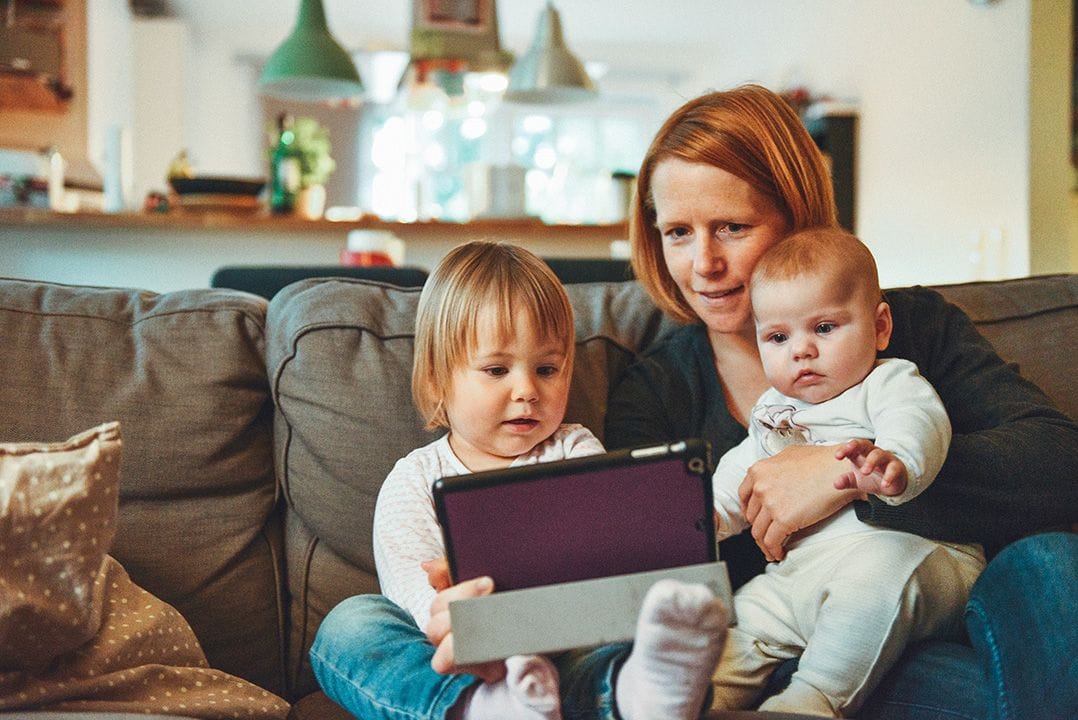 An infant, toddler, and woman sitting on a couch looking at a tablet 